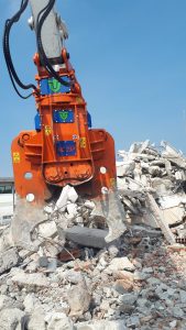 An orange Trevi Benne primary pulveriser excavator attachment hovers over concrete rubble and rebar scrap for demolition.